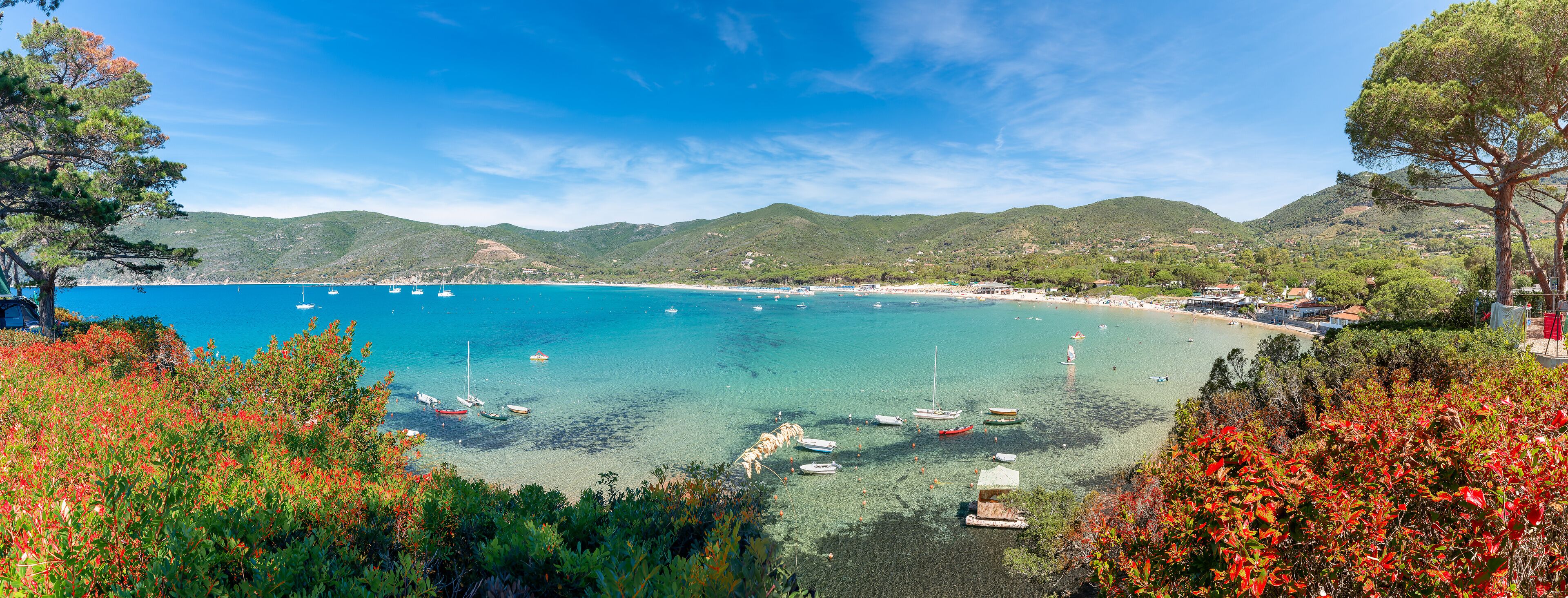 A coastal landscape with clear turquoise waters, boats, and a backdrop of green hills.