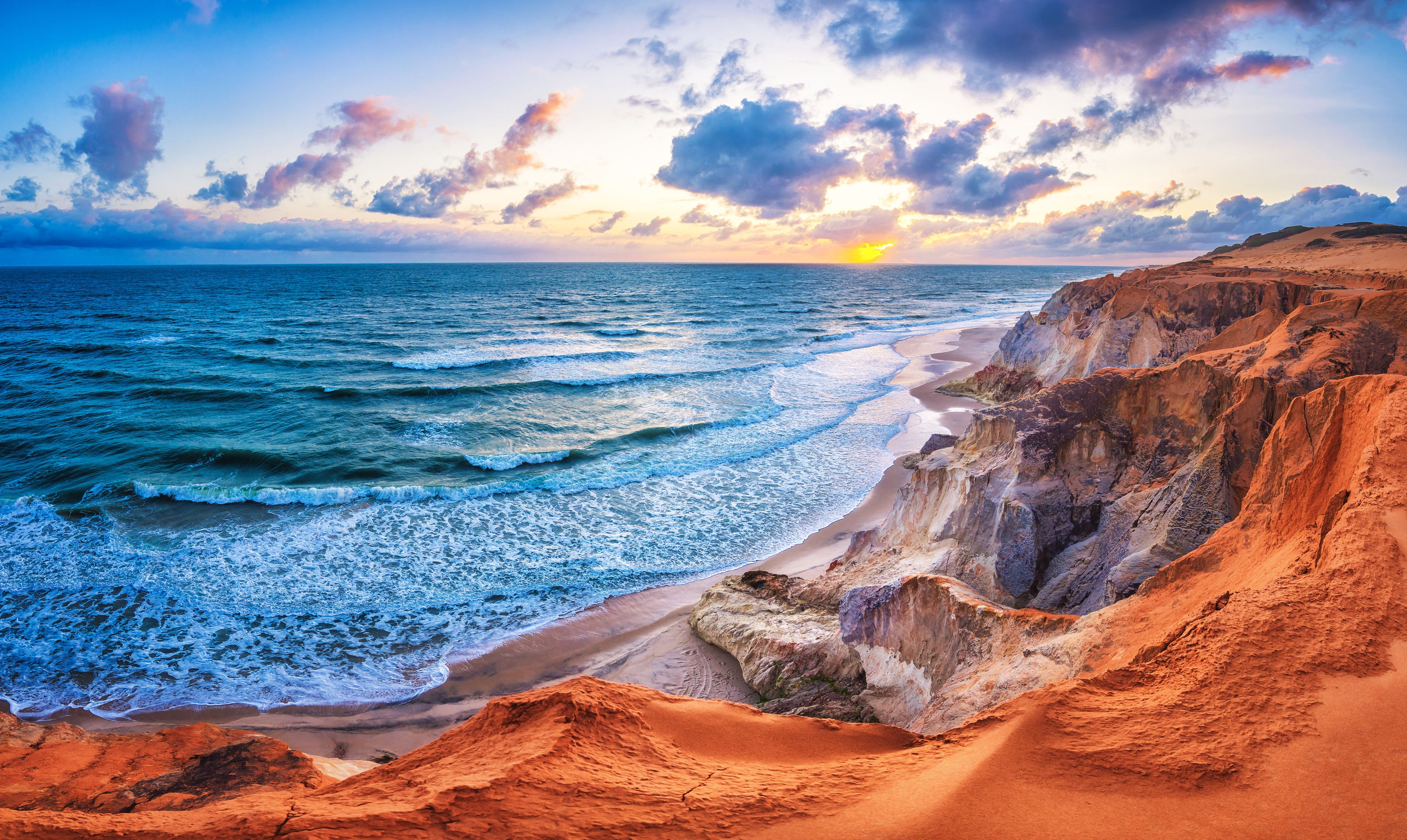 Paisagem costeira com ondas quebrando em uma praia arenosa, penhascos e um pôr do sol ao fundo.