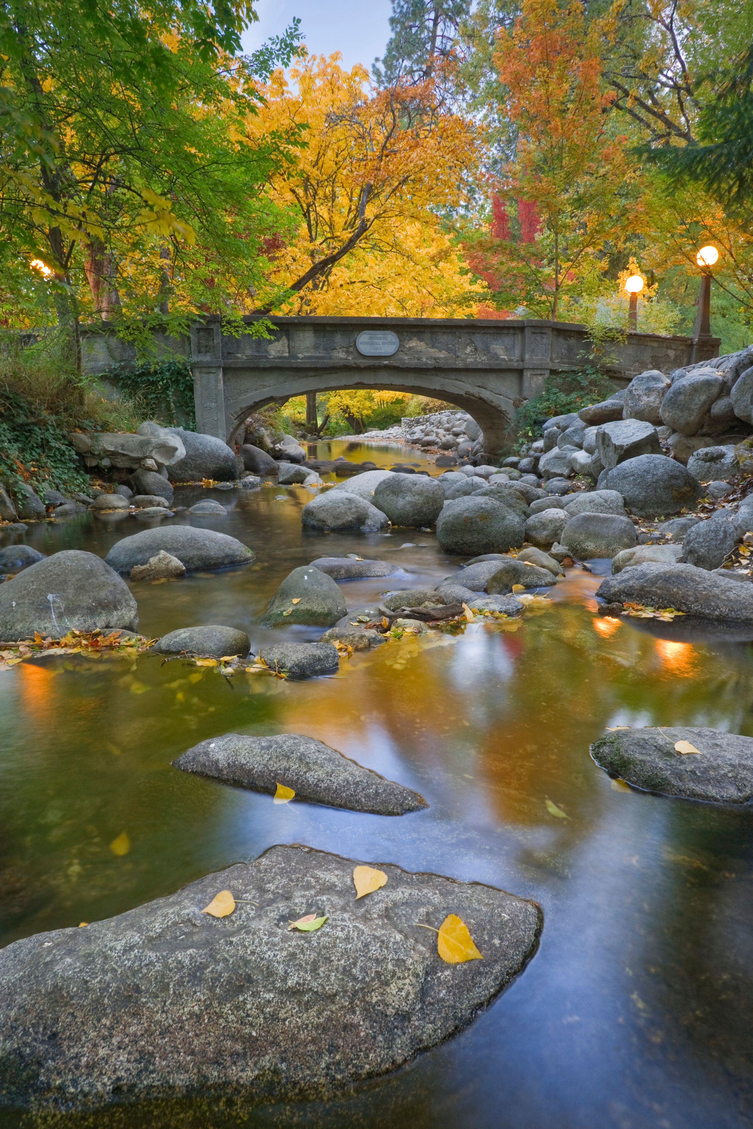 A stone bridge over a calm stream with autumn leaves and rocks.