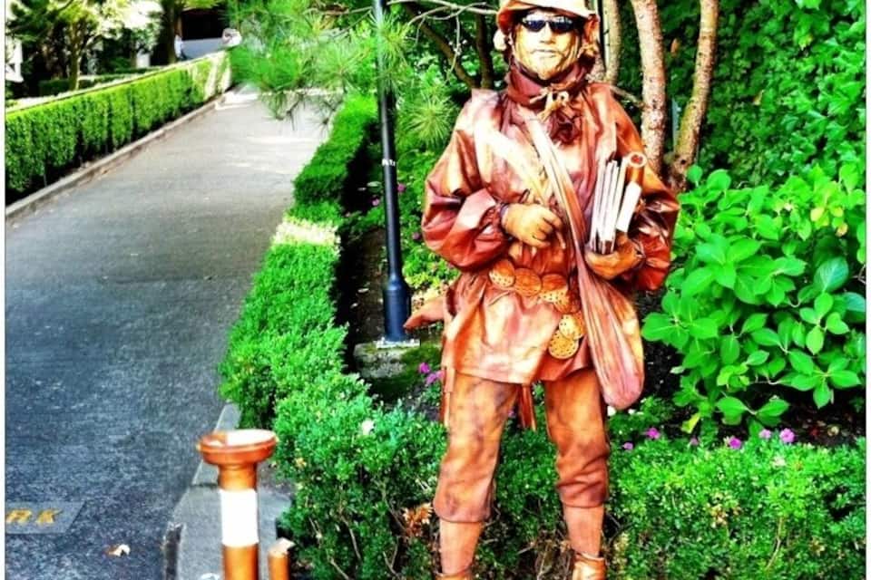 En staty av en person klädd i en brun outfit, med hatt och solglasögon, som håller i en pipa och en bok och står på en piedestal i en park med gröna buskar och träd.
