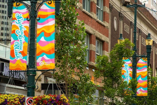 Bunte Banner hängen an einem Straßenlaternenpfahl, ein Blumenkorb und ein Gebäude mit der Aufschrift 'downtown calgary'.