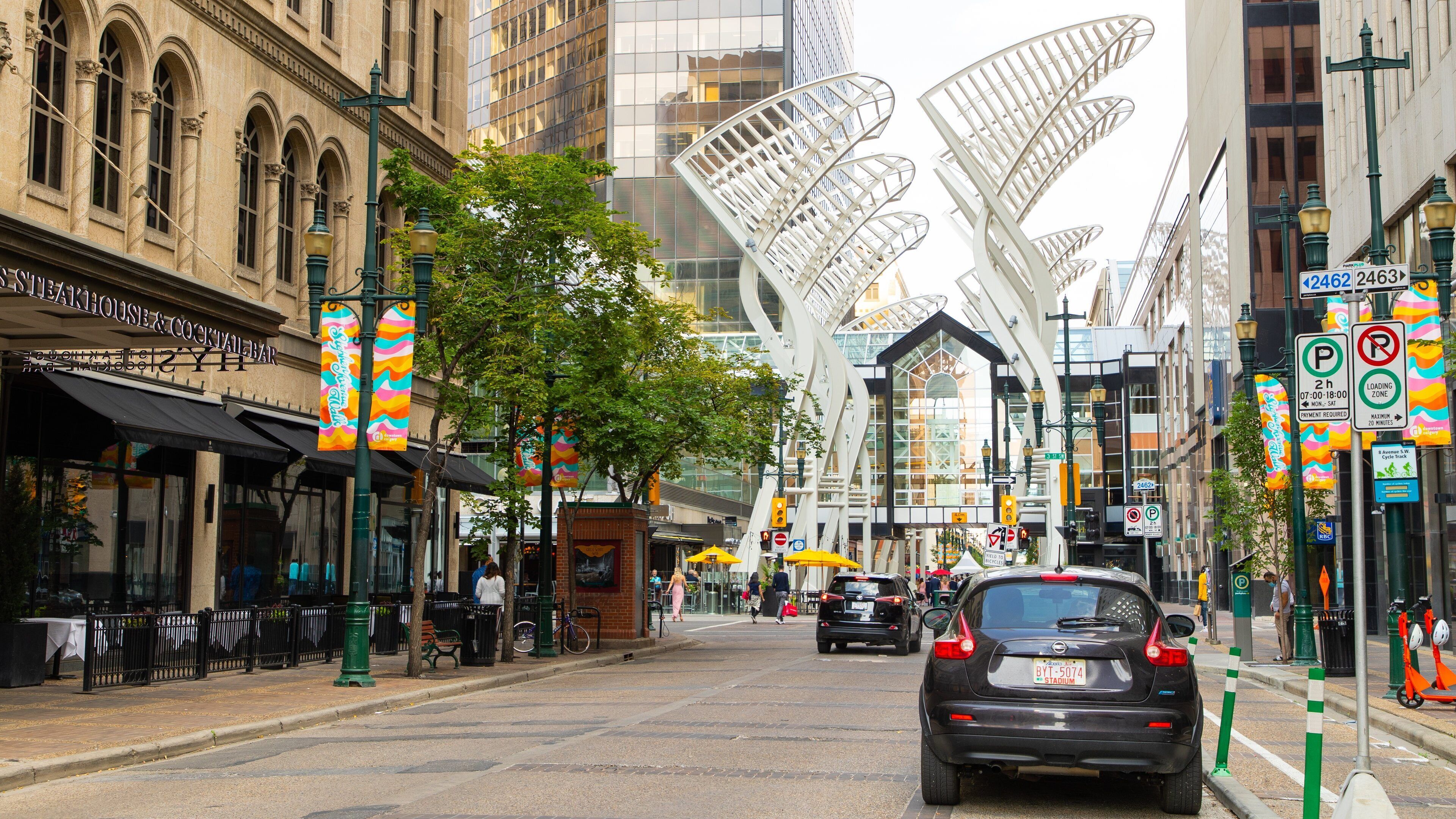 A city street with a modern building featuring unique white architecture. There are street signs, a car, and pedestrians.