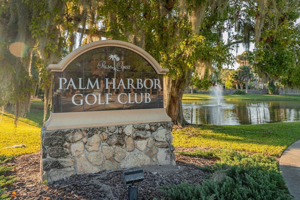 A sign for Palm Harbor Golf Club with a pond and fountain in the background.