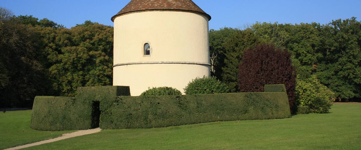 A round tower with a conical roof, surrounded by neatly trimmed hedges and trees, set against a clear blue sky.