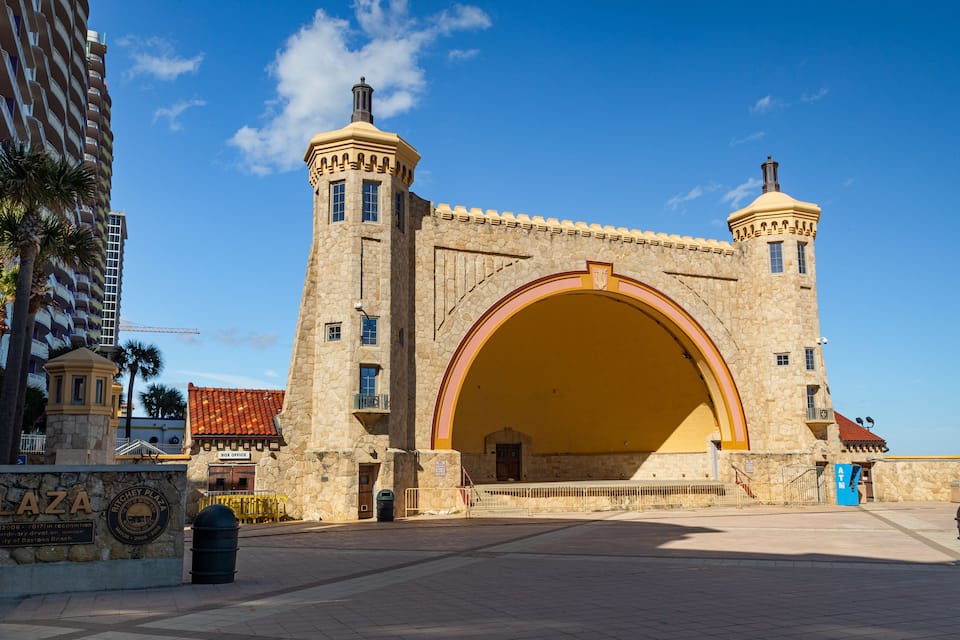 Un edificio histórico de piedra con una gran entrada arqueada, palmeras y un cielo azul despejado.