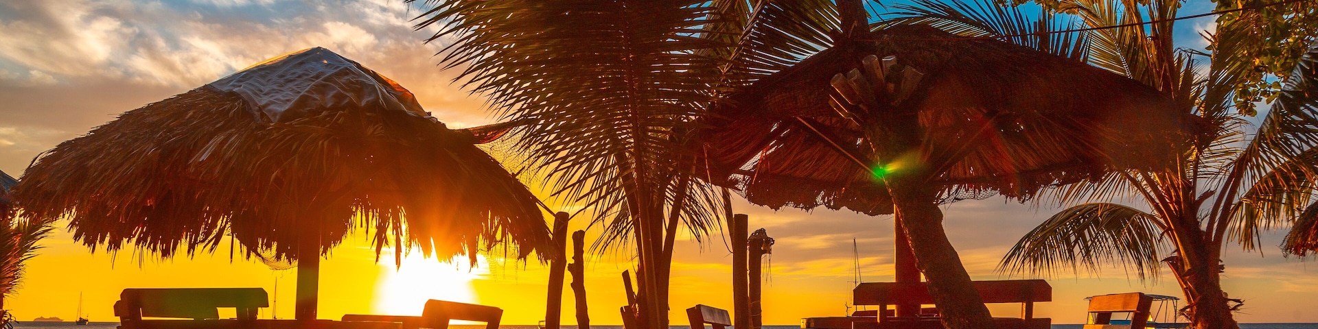 Uma praia com bancos de madeira e um guarda-sol de palha, palmeiras e o oceano ao pôr do sol.