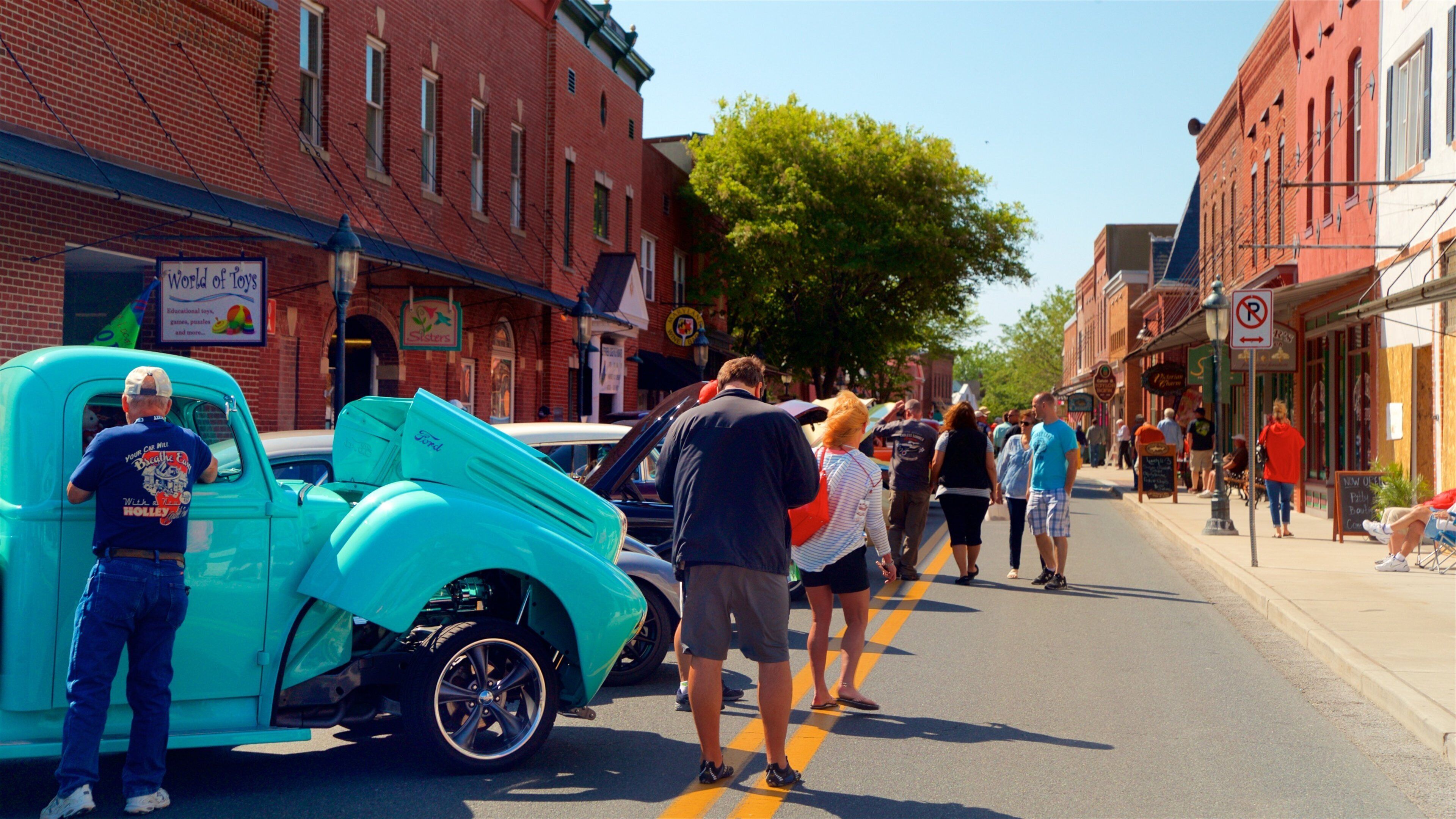 A turquoise vintage car parked on a street lined with brick buildings, a 'World of Toys' sign, and a 'No Parking' sign.
