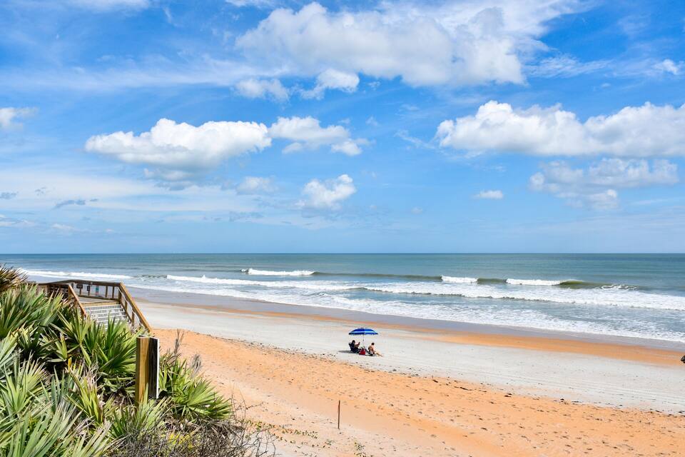 Een strand met een houten trap naar het zand, een persoon die onder een blauwe parasol zit, en de oceaan op de achtergrond.