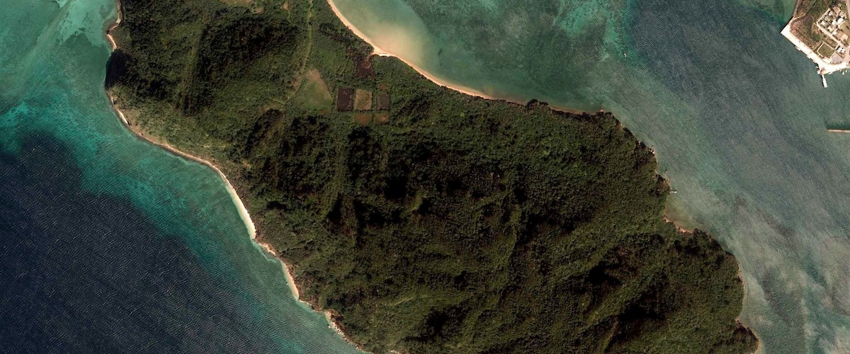 An aerial view of a tropical island with clear turquoise waters, dense green vegetation, and a small settlement.