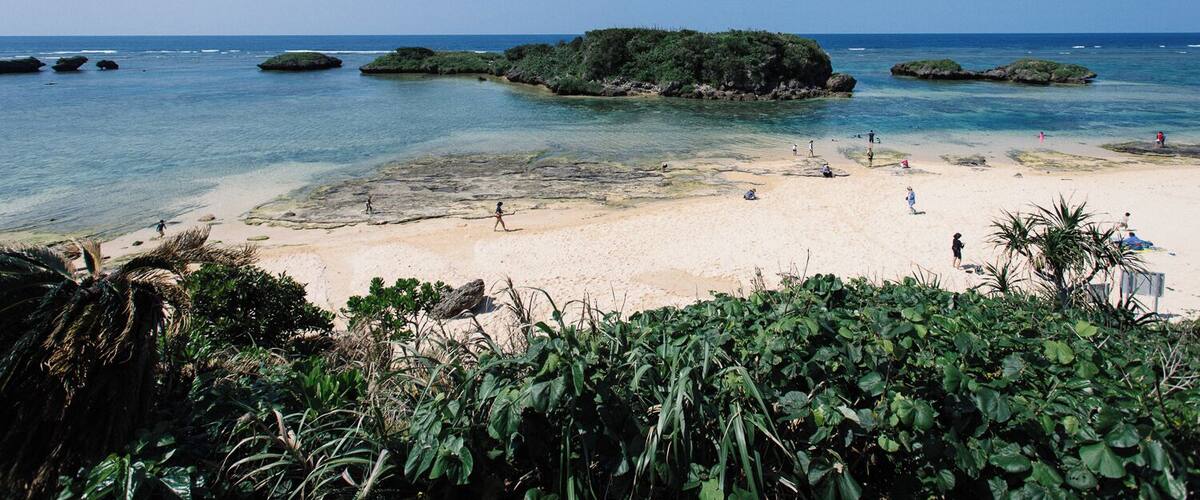 A beach with clear turquoise water, rocky outcrops, and lush greenery in the foreground.