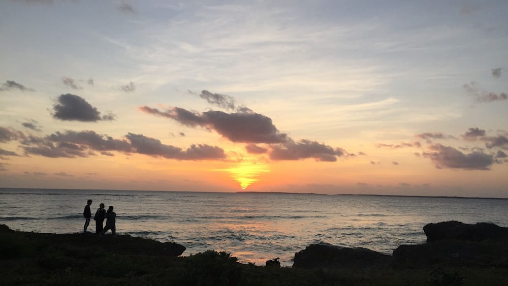 A sunset over the ocean with silhouettes of people on a rocky shore.