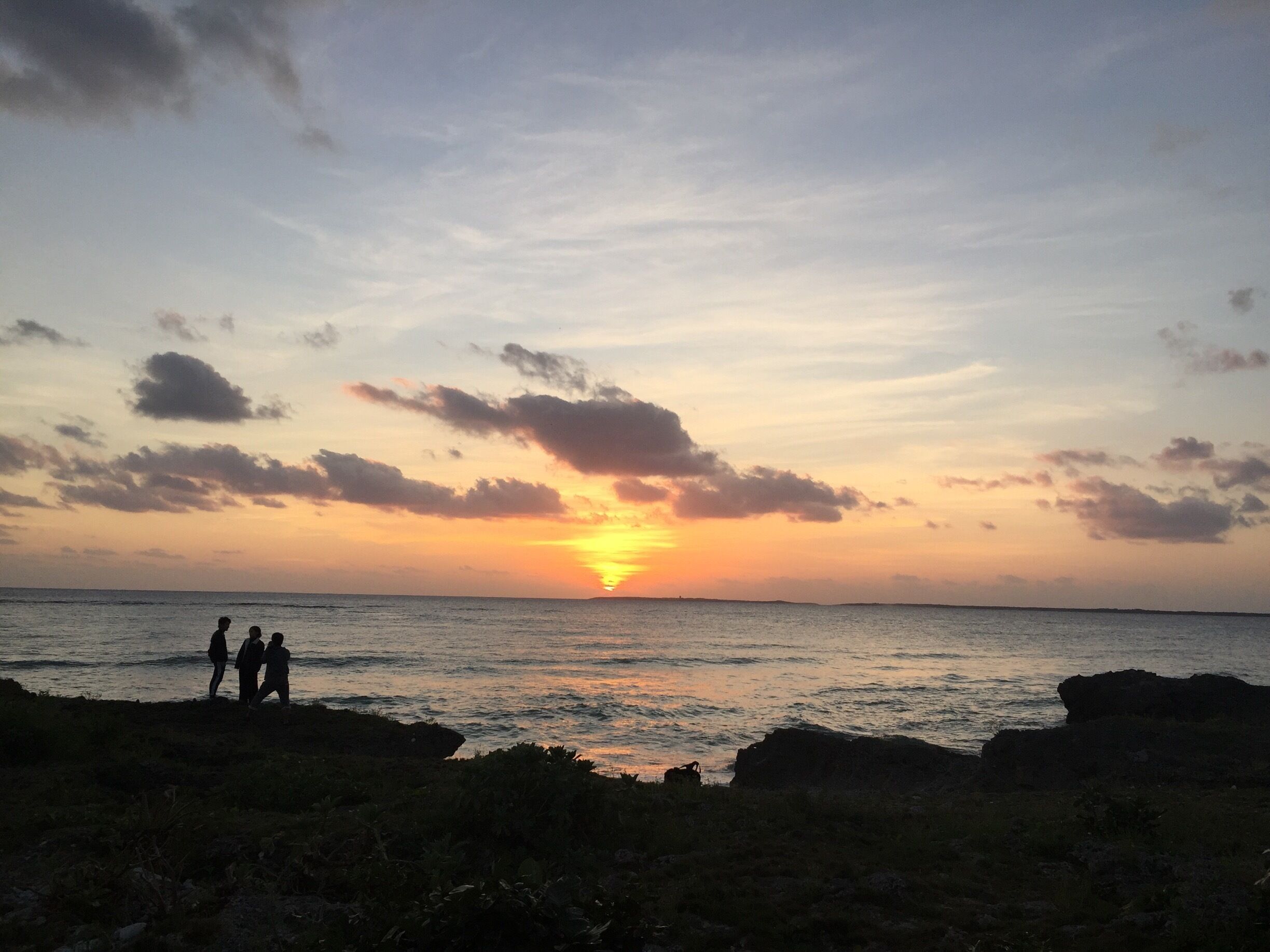 A sunset over the ocean with silhouettes of people on a rocky shore.