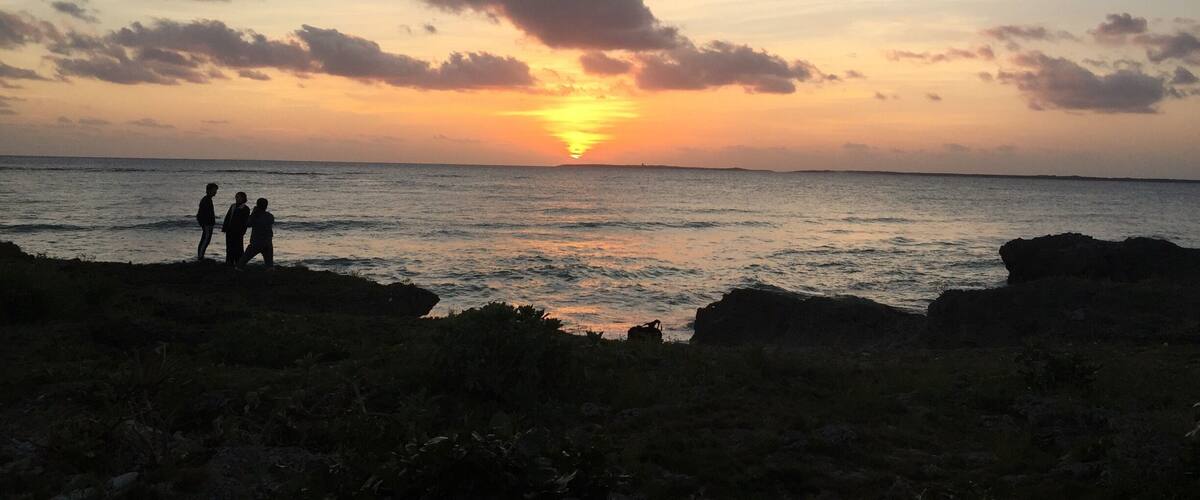 A sunset over the ocean with silhouettes of people on a rocky shore.