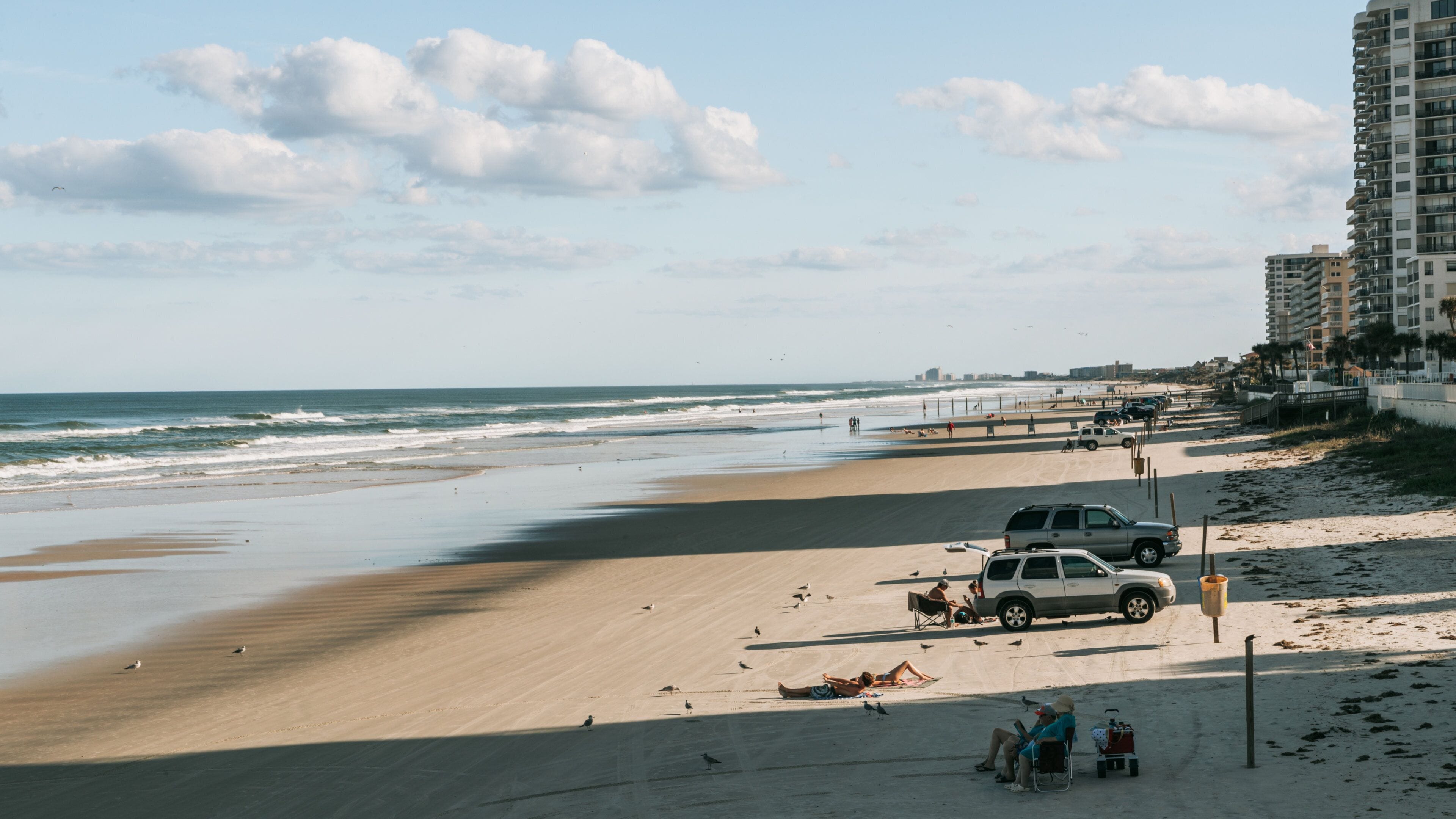A beach with a sandy shore, waves, and a few people. There are buildings in the background and a parked car near the water.