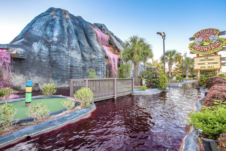 A water-themed attraction with a rocky backdrop, a wooden bridge, and a sign for Molten Mountain Golf.