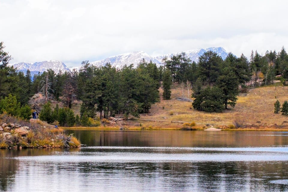 A calm lake surrounded by dense forests and snow-capped mountains in the background.