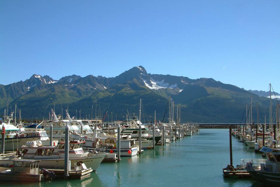 A marina with boats docked in calm waters, surrounded by mountains with snow-capped peaks.