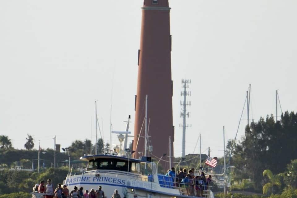 A boat named 'PASTIME PRINCESS' with passengers, a lighthouse in the background, and a clear sky.