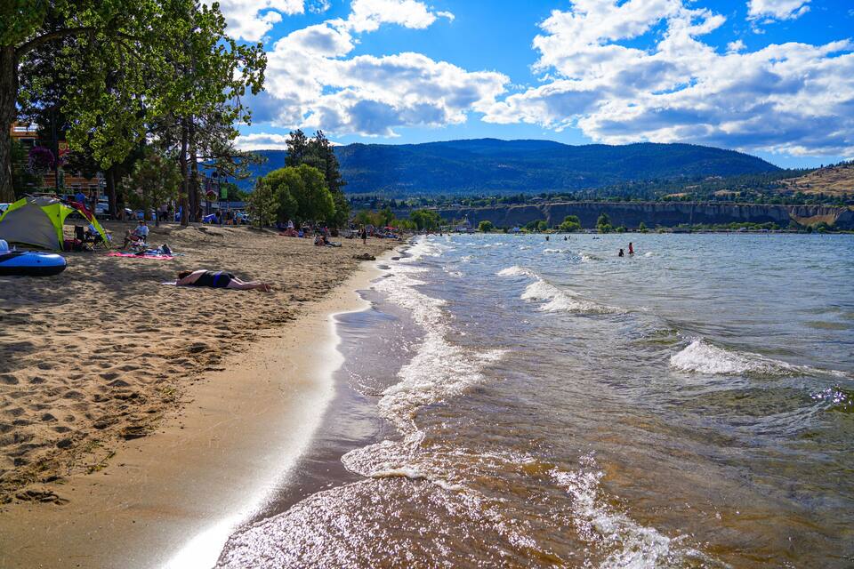 Una playa arenosa con tiendas de campaña, gente y un lago con montañas al fondo.