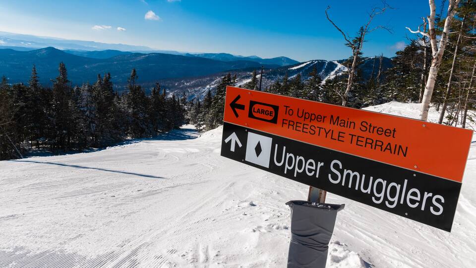 A snowy mountain landscape with a sign indicating directions to Upper Main Street and Upper Smugglers.