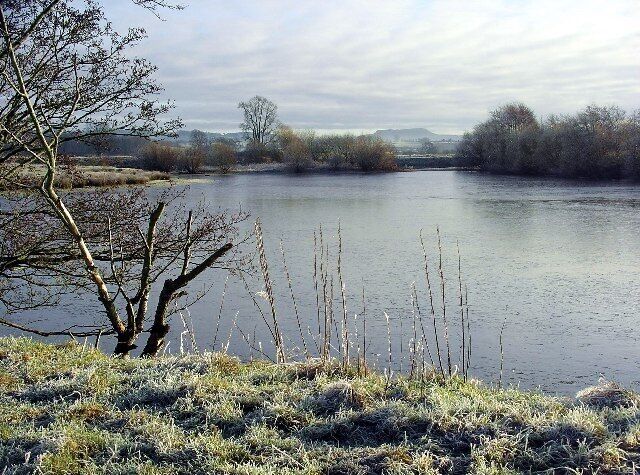 A calm river with frost-covered grass and leafless trees in the foreground.