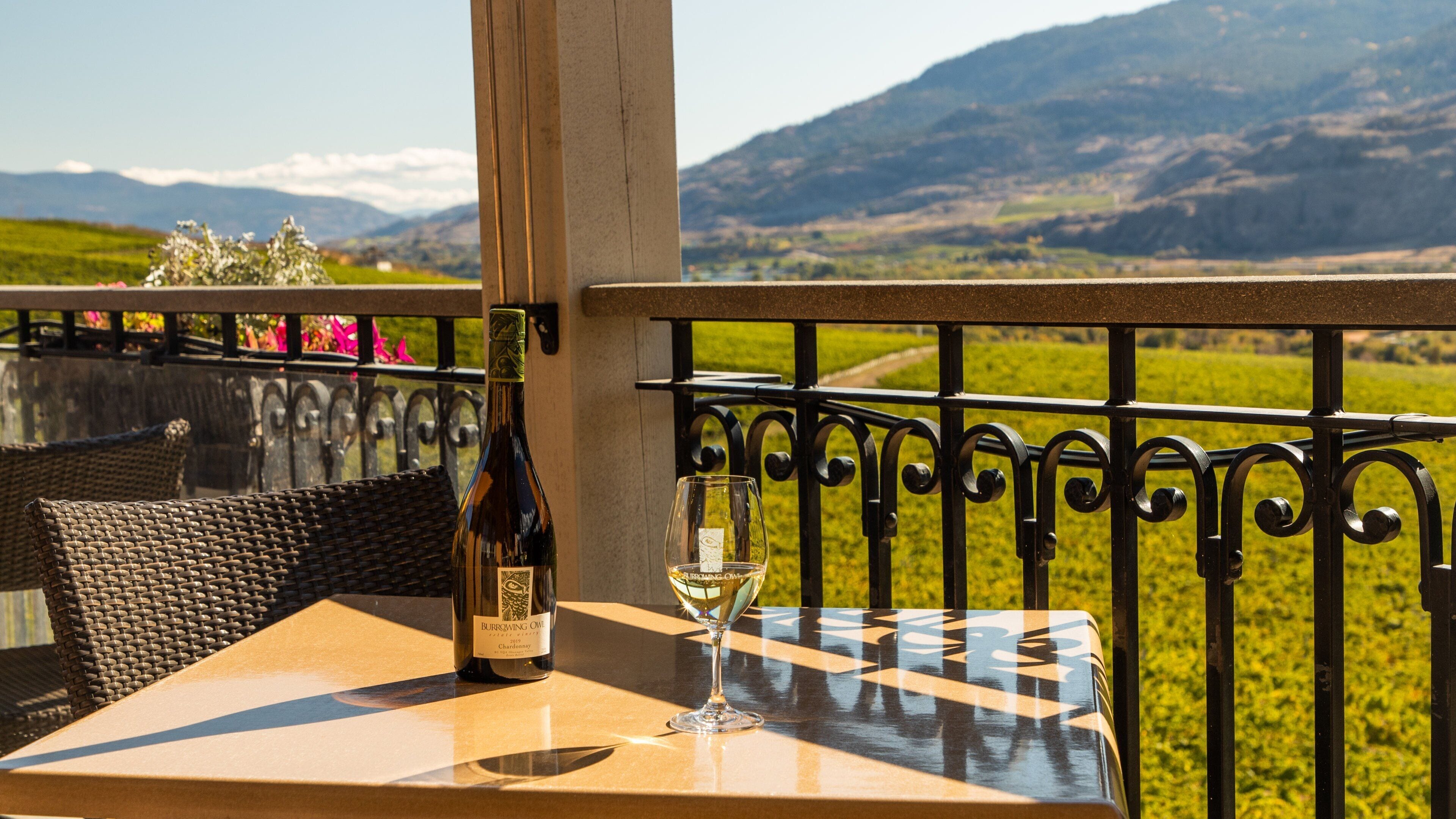 A bottle of Burrowing Owl Chardonnay and a glass on a table with a view of a mountainous landscape.