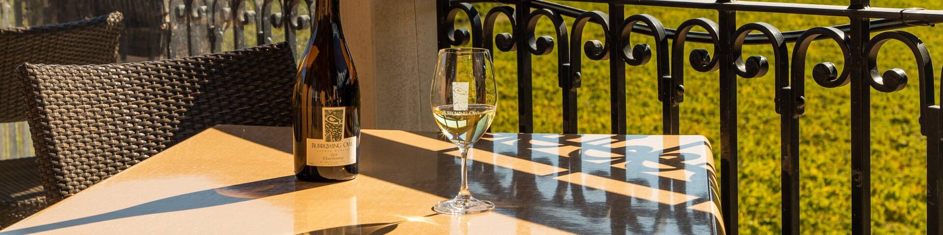 A bottle of Burrowing Owl Chardonnay and a glass on a table with a view of a mountainous landscape.