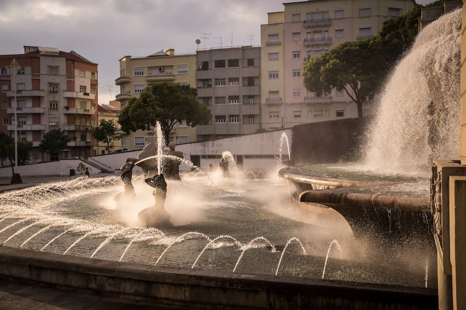 A fountain with water jets in a public square, surrounded by multi-story buildings and trees.