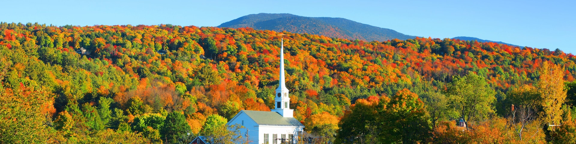 A white church with a steeple, surrounded by autumn-colored trees and a mountain in the background.