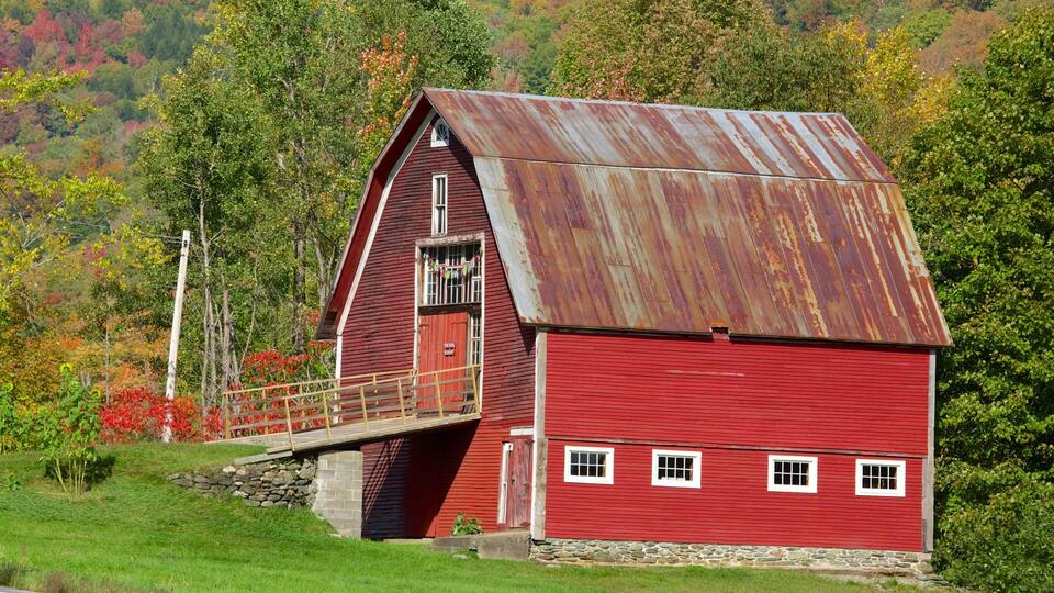 A red barn with a metal roof, surrounded by green trees and a stone wall.
