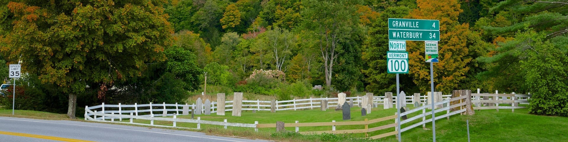 A scenic road with a white fence and gravestones, surrounded by autumn-colored trees and a hill in the background.