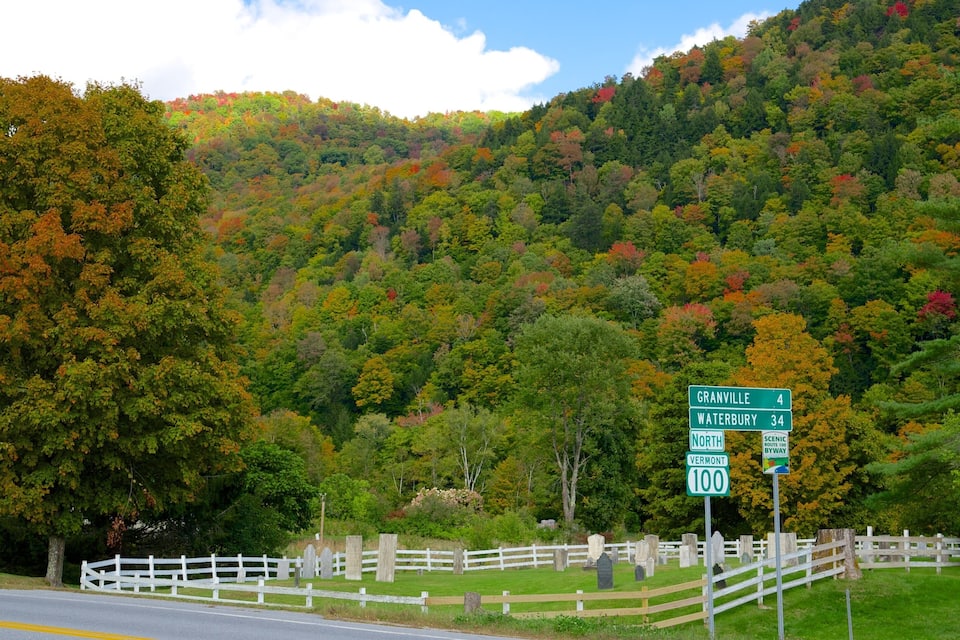 A scenic road with a white fence and gravestones, surrounded by autumn-colored trees and a hill in the background.