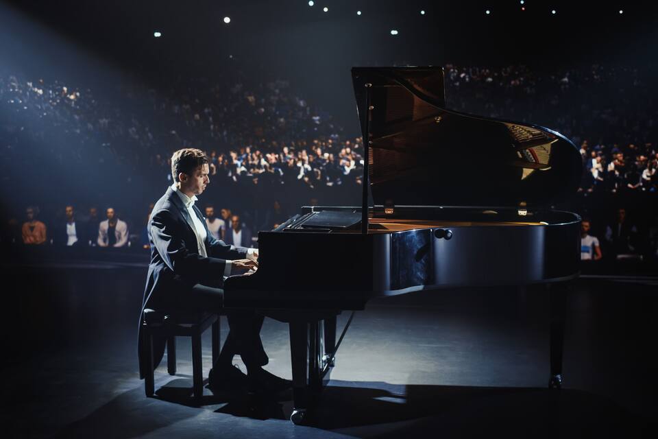 Un pianista tocant un piano de cua en una sala de concerts poc il·luminada, amb un públic al fons.