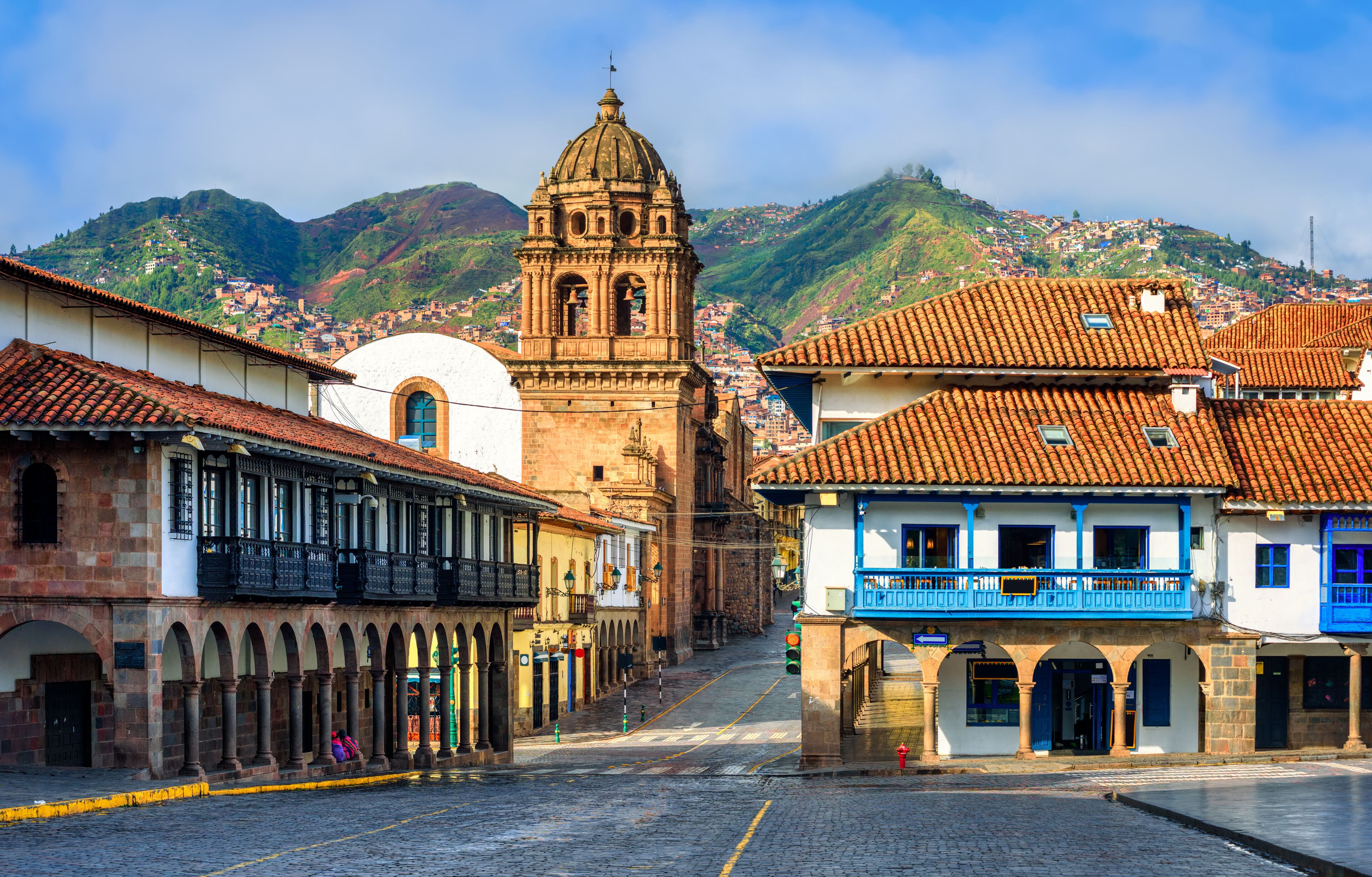 A cobblestone street lined with historic buildings, a prominent church with a bell tower, and a mountainous backdrop.