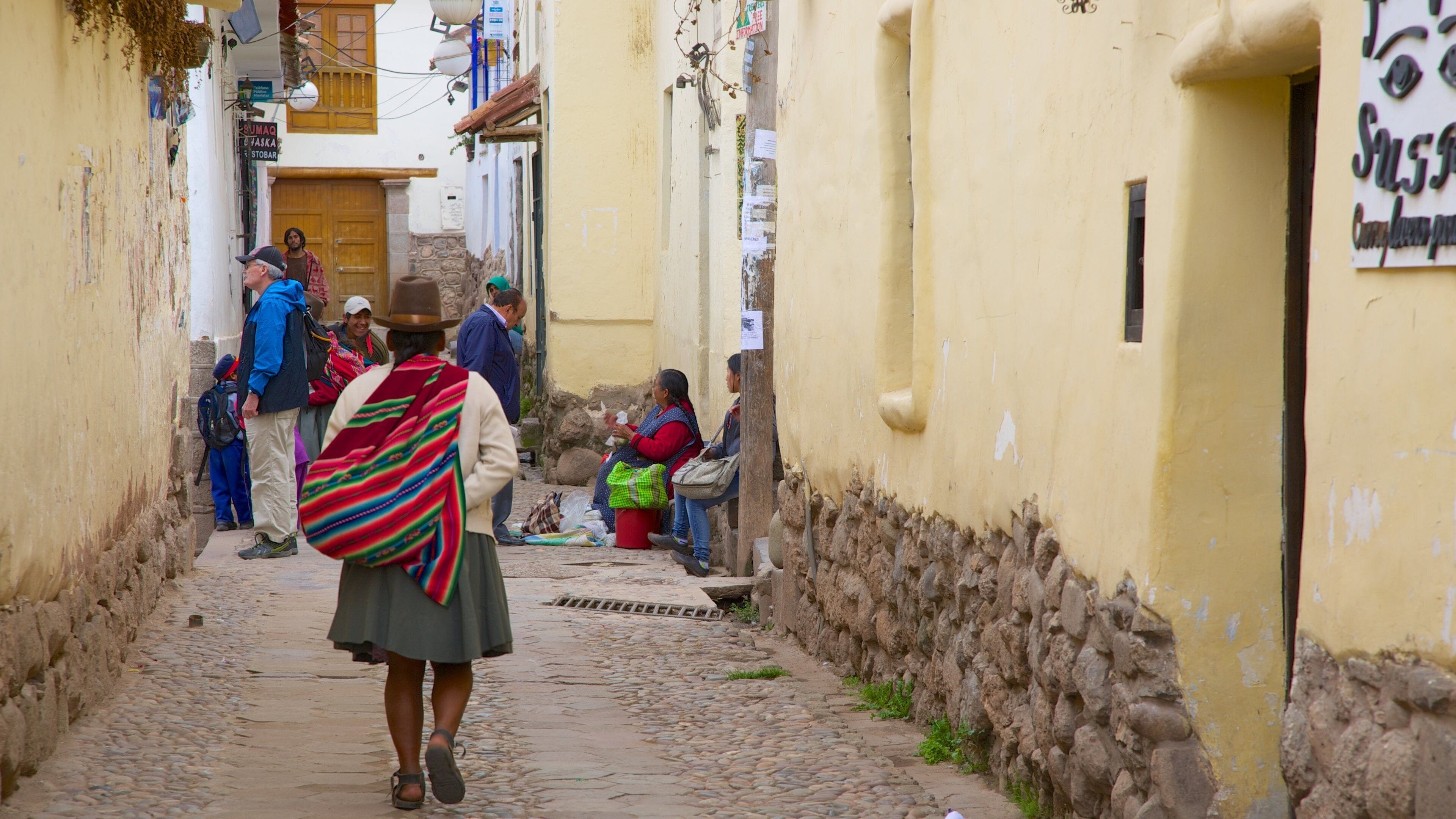 A narrow cobblestone street lined with yellow buildings, a person with a colorful shawl walking down the path, and a few people sitting on the ground.