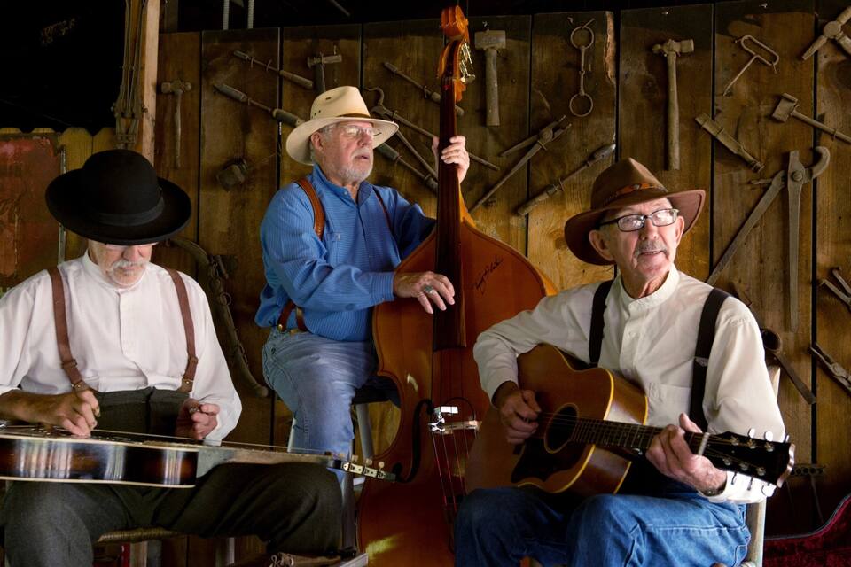 Three men playing string instruments in a rustic setting with wooden walls and tools.