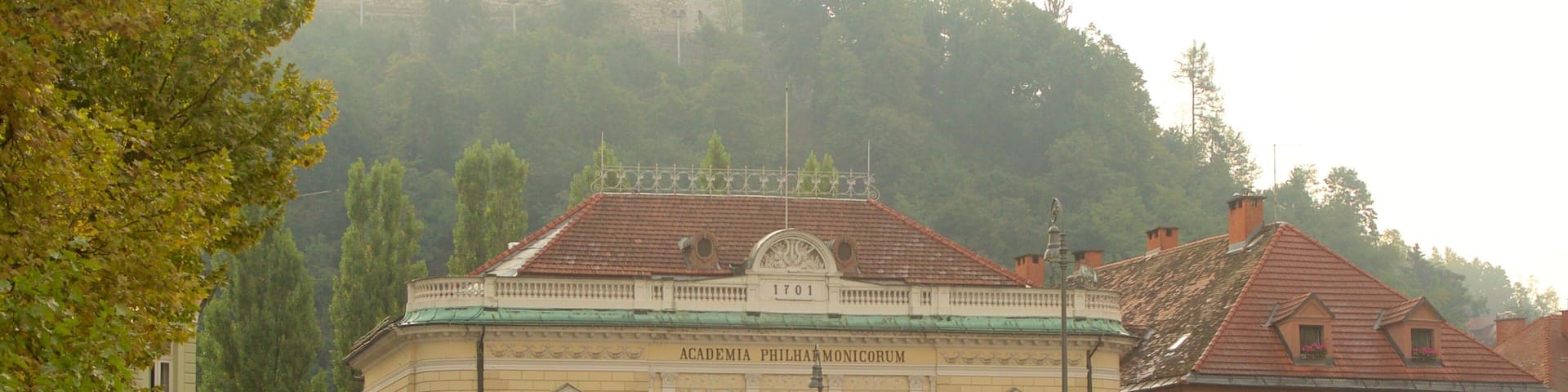 A historic building with the inscription "Academia Philharmonica Monorum" and a red-roofed building adjacent to it.