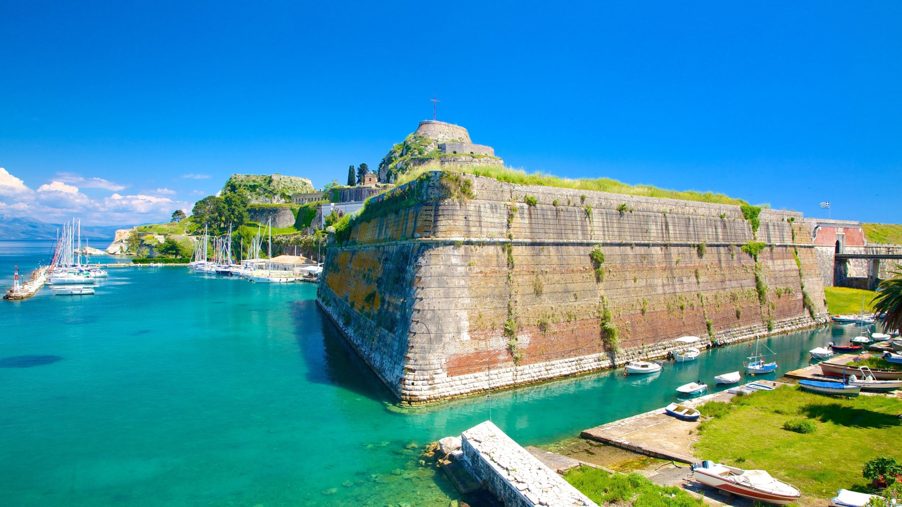 A coastal fortification with a cross on top, surrounded by clear turquoise waters and a marina with boats.