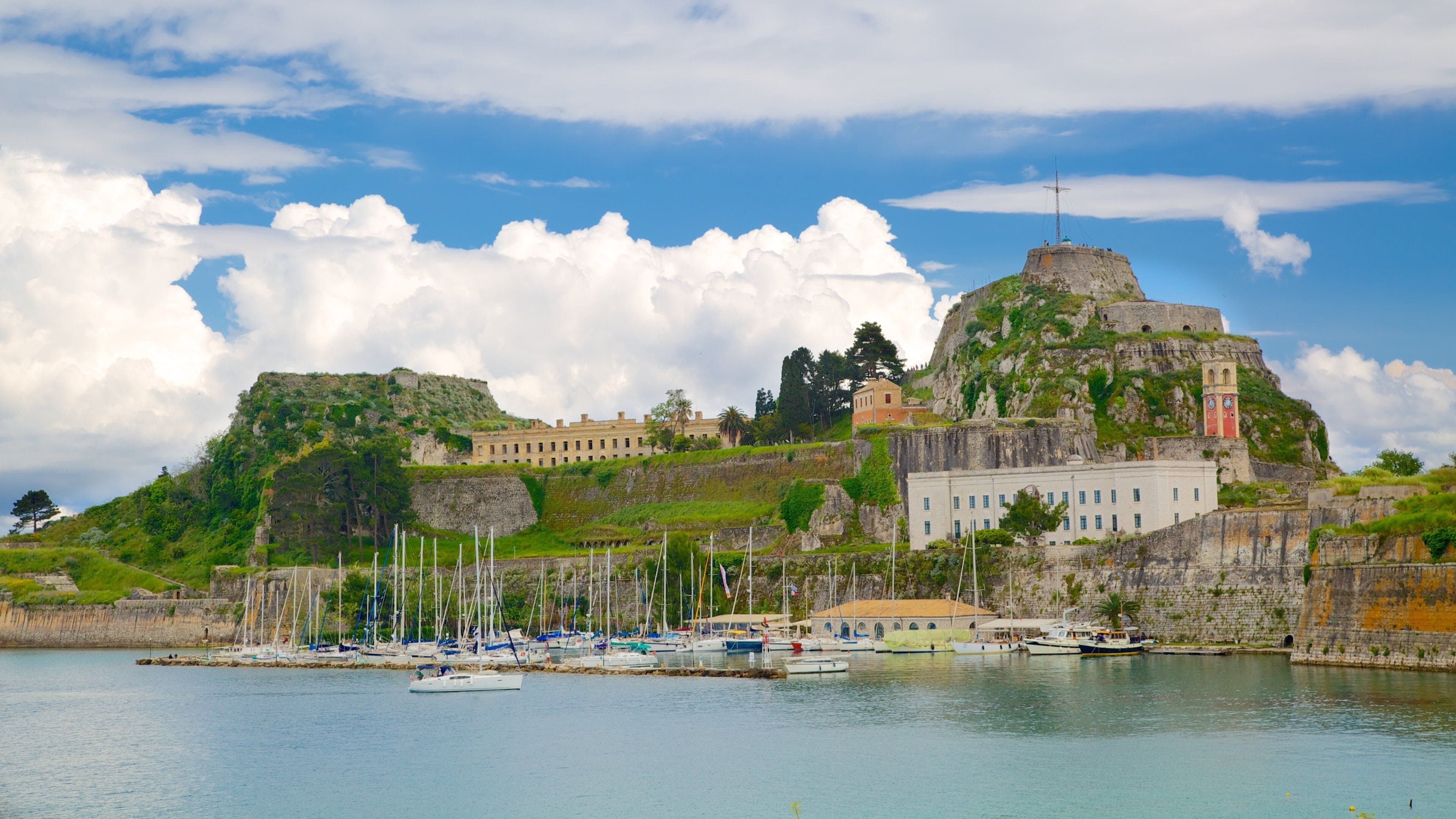 Une ville côtière avec un château imposant perché sur une colline, un port de plaisance rempli de bateaux et un ciel d’un bleu limpide.