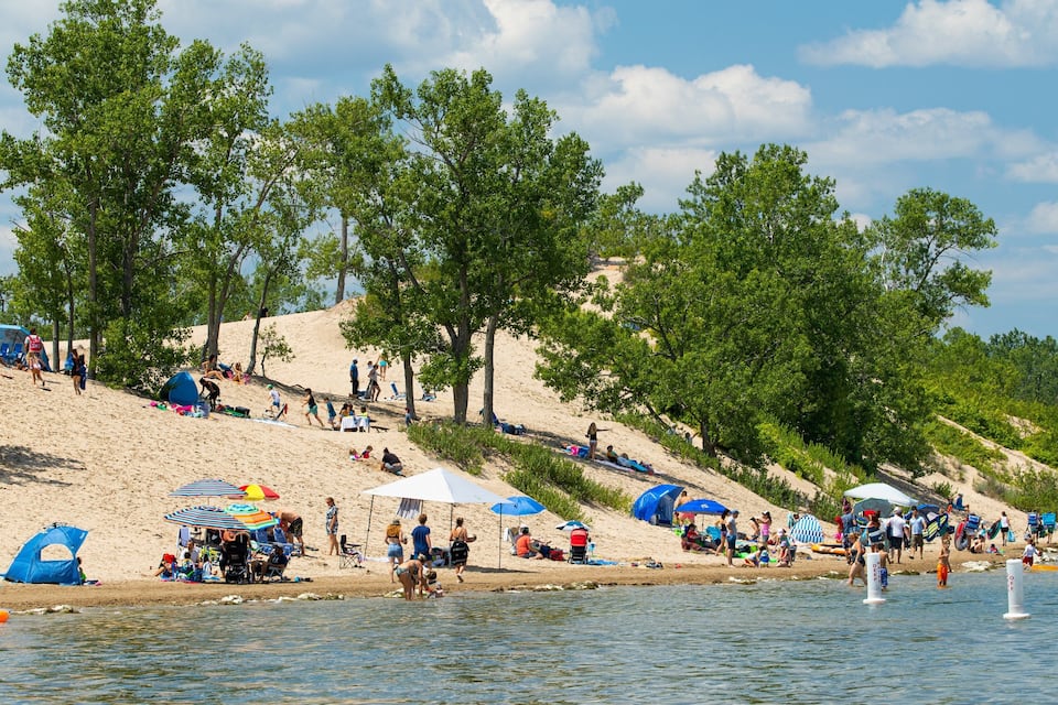 Una spiaggia con persone che prendono il sole e nuotano, circondata da alberi e dune di sabbia.