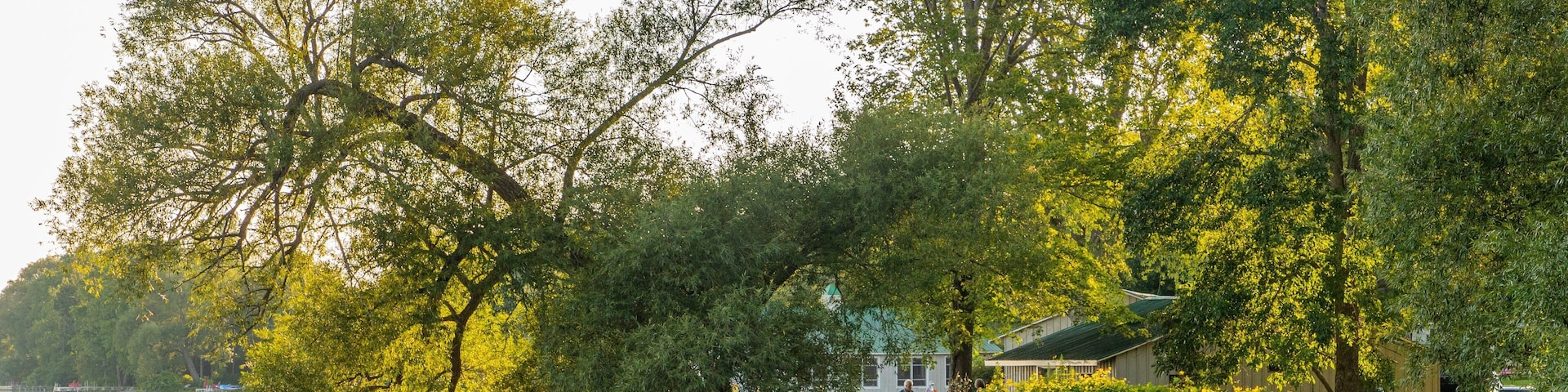 A calm lake with trees reflecting sunlight, a small boat, and a house with a green roof by the water's edge.