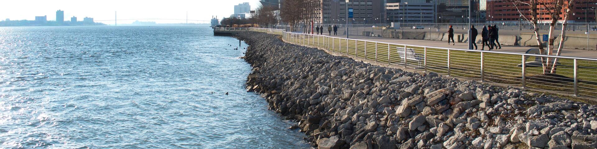 A waterfront cityscape with a rocky breakwater, a pedestrian walkway, and modern high-rise buildings.