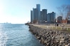 A waterfront cityscape with a rocky breakwater, a pedestrian walkway, and modern high-rise buildings.