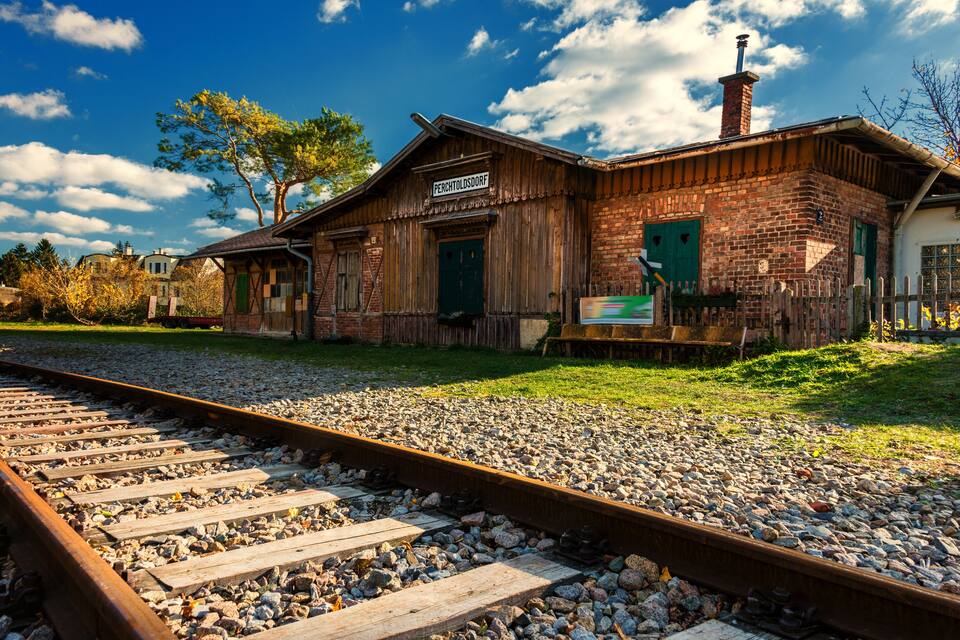 A rustic train station with a wooden building labeled "PERCHTOLDSDORF" and a railway track in the foreground.