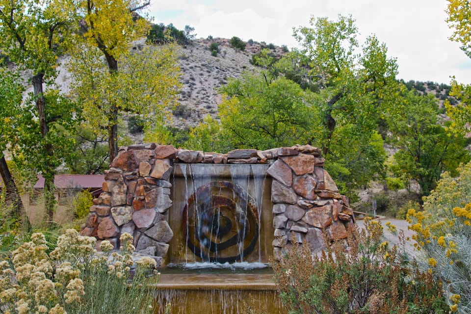 A rustic stone waterfall in a natural setting with surrounding vegetation and a hillside in the background.