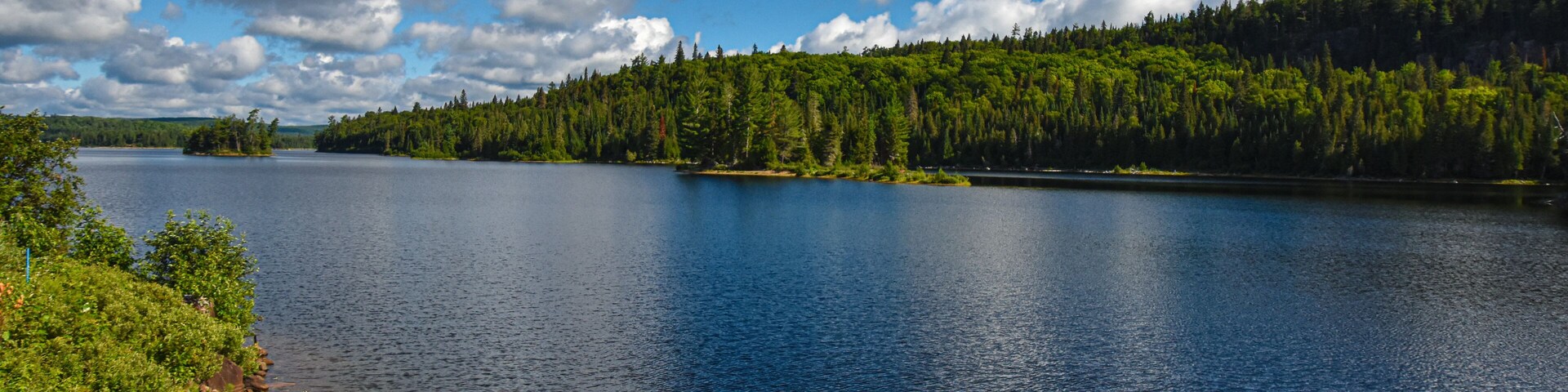 Un lago tranquilo con un pequeño bote azul en la orilla, rodeado de un bosque frondoso y un cielo azul despejado con nubes.