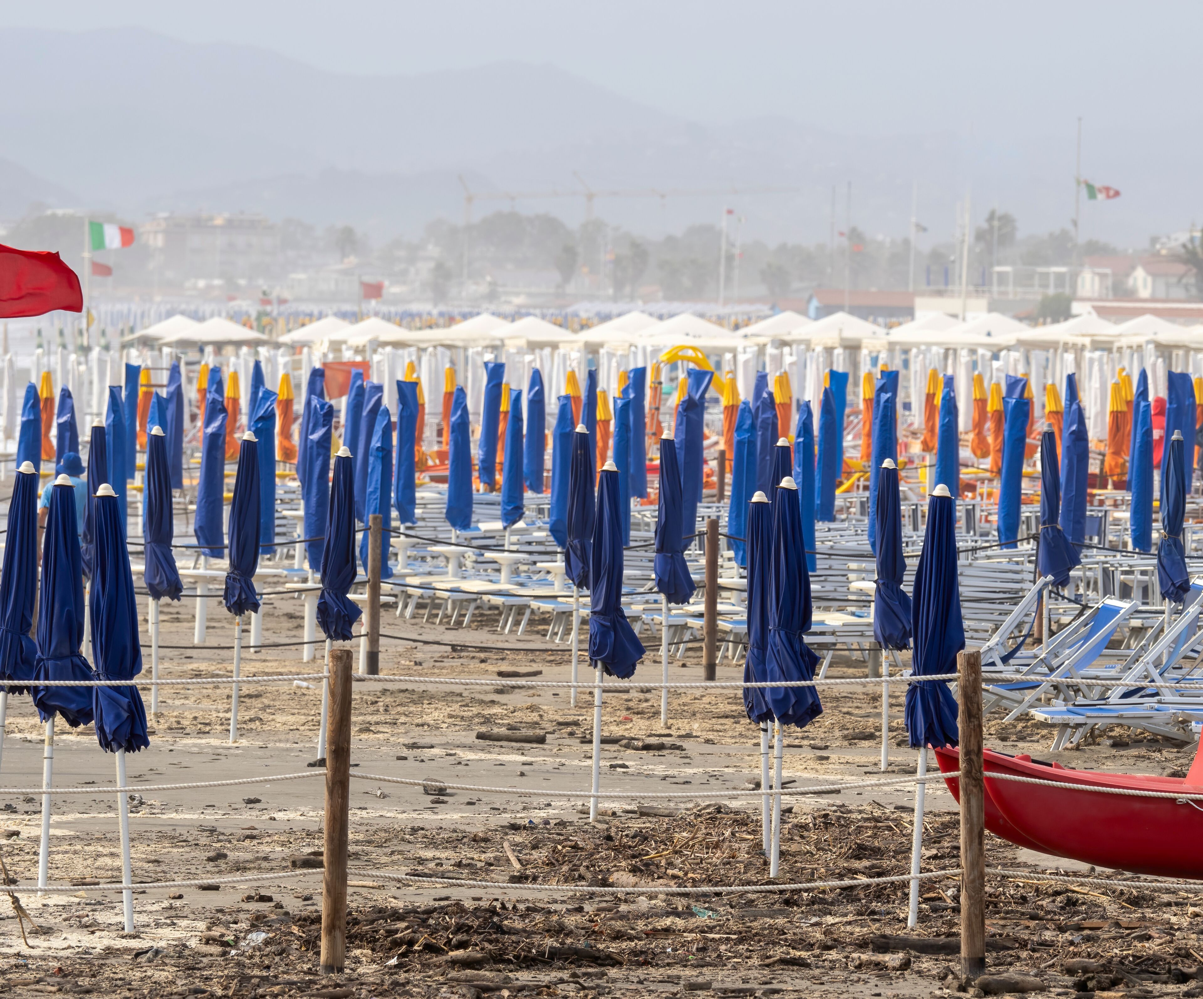 Rows of beach umbrellas and chairs on a sandy beach with a red boat in the foreground.