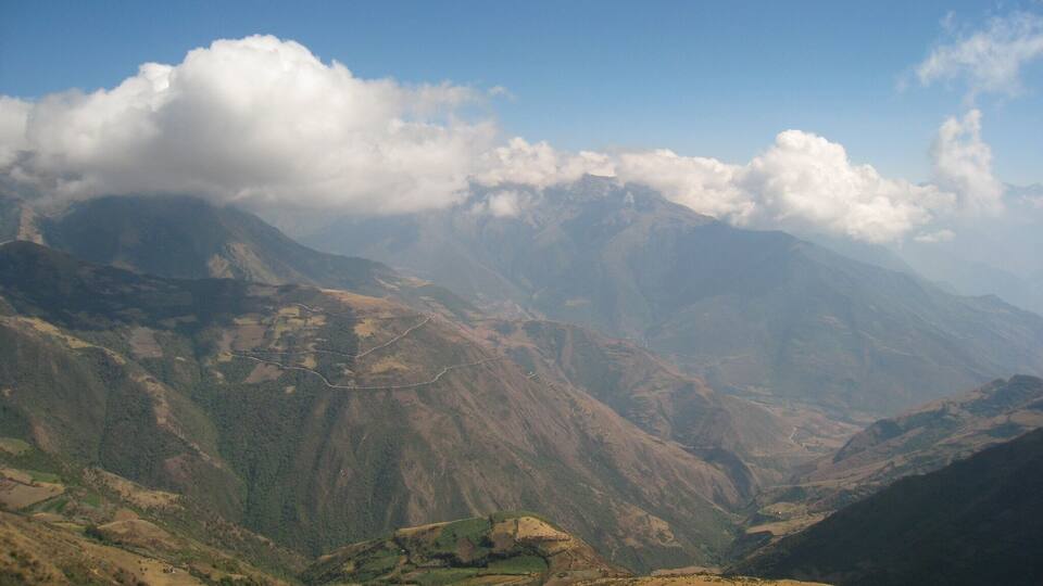 A mountainous landscape with a winding road, green valleys, and distant peaks under a partly cloudy sky.
