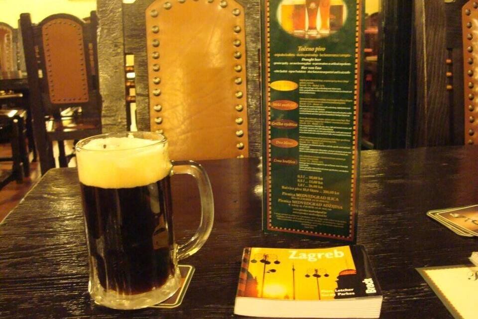 A dark beer in a glass on a wooden table with a menu and a brochure titled "Zagreb".