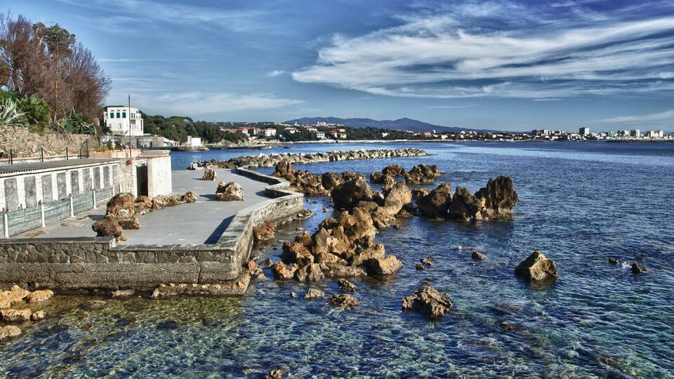 A rocky shoreline with clear water, a promenade, and distant cityscape.
